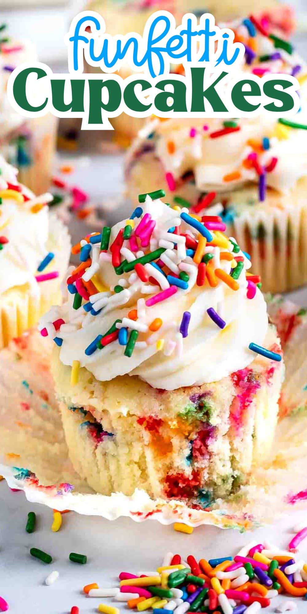 A funfetti cupcake with white frosting and colorful sprinkles sits on a light surface, surrounded by more cupcakes. The vibrant sprinkles add a festive touch. The partially unwrapped cupcake reveals a colorful, speckled interior.