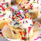 A vanilla cupcake topped with white frosting and colorful sprinkles, with its wrapper partially peeled away. More similarly decorated cupcakes are in the background on a white surface.