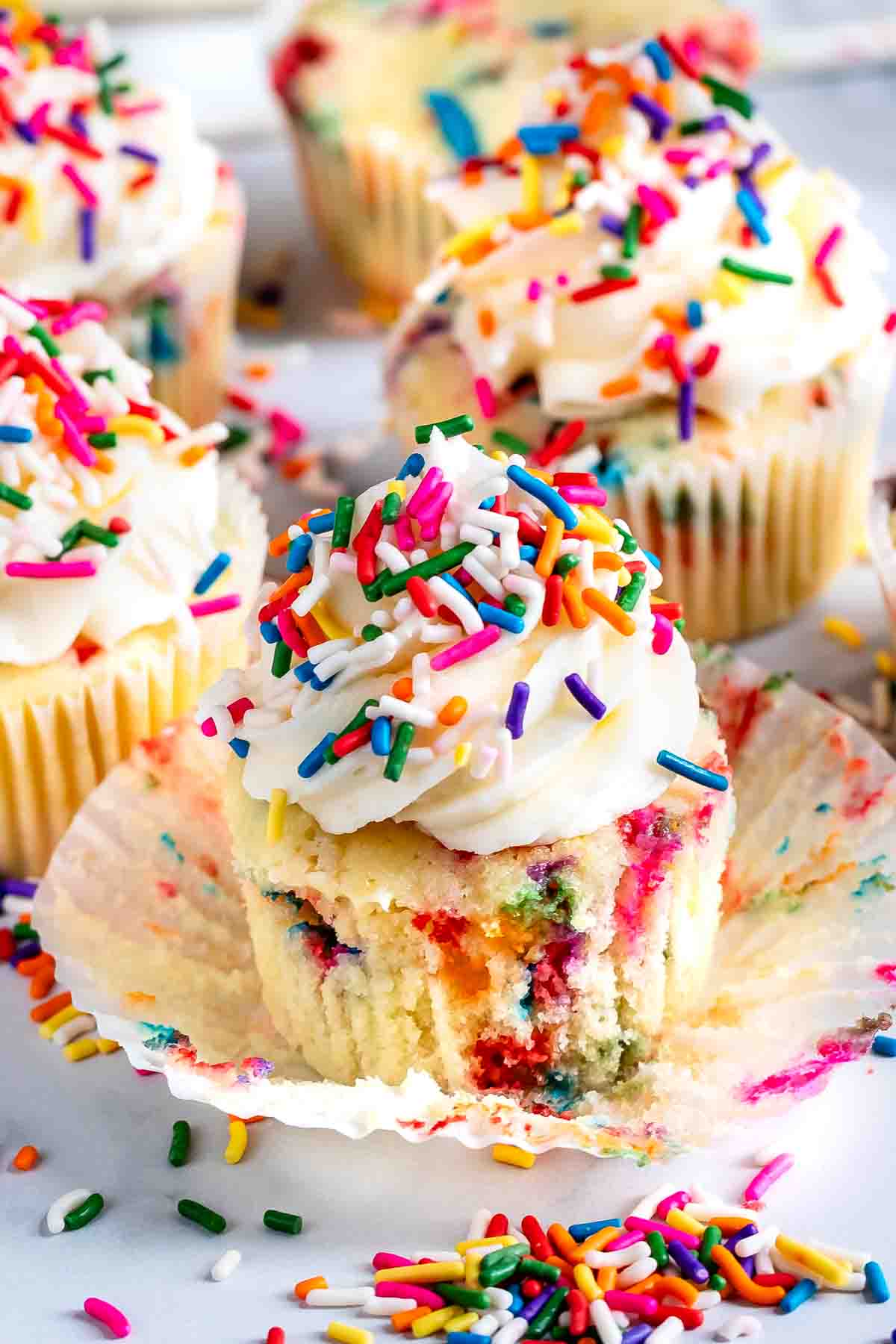 A vanilla cupcake topped with white frosting and colorful sprinkles, with its wrapper partially peeled away. More similarly decorated cupcakes are in the background on a white surface.
