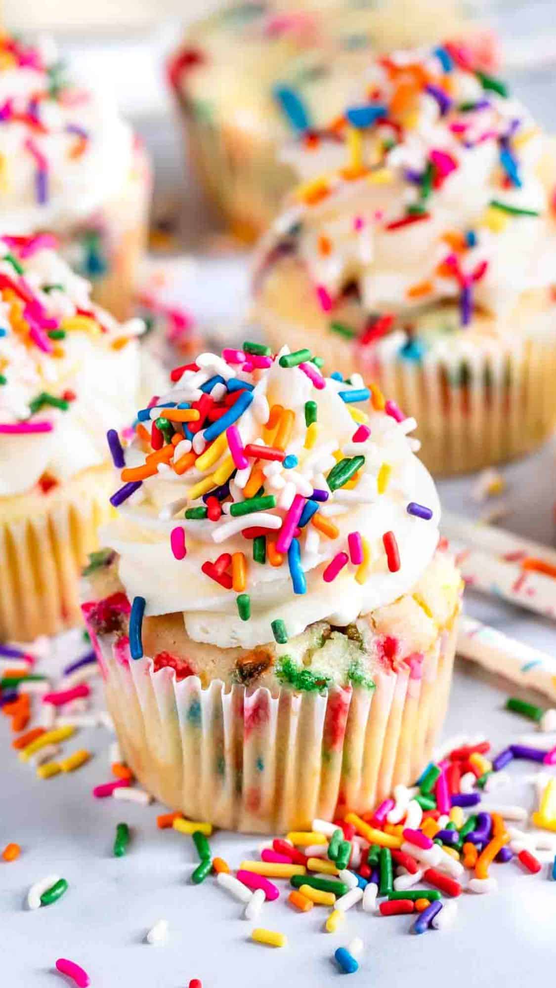 Cupcakes topped with white frosting and colorful sprinkles are displayed. Some sprinkles have scattered on the surface around them. The cupcakes have a festive appearance with bursts of color in their batter.