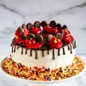 A white frosted cake with a chocolate drip, decorated with red and yellow icing, chocolate pieces, and colorful sprinkles on the base, displayed on a white cake stand against a marble background.