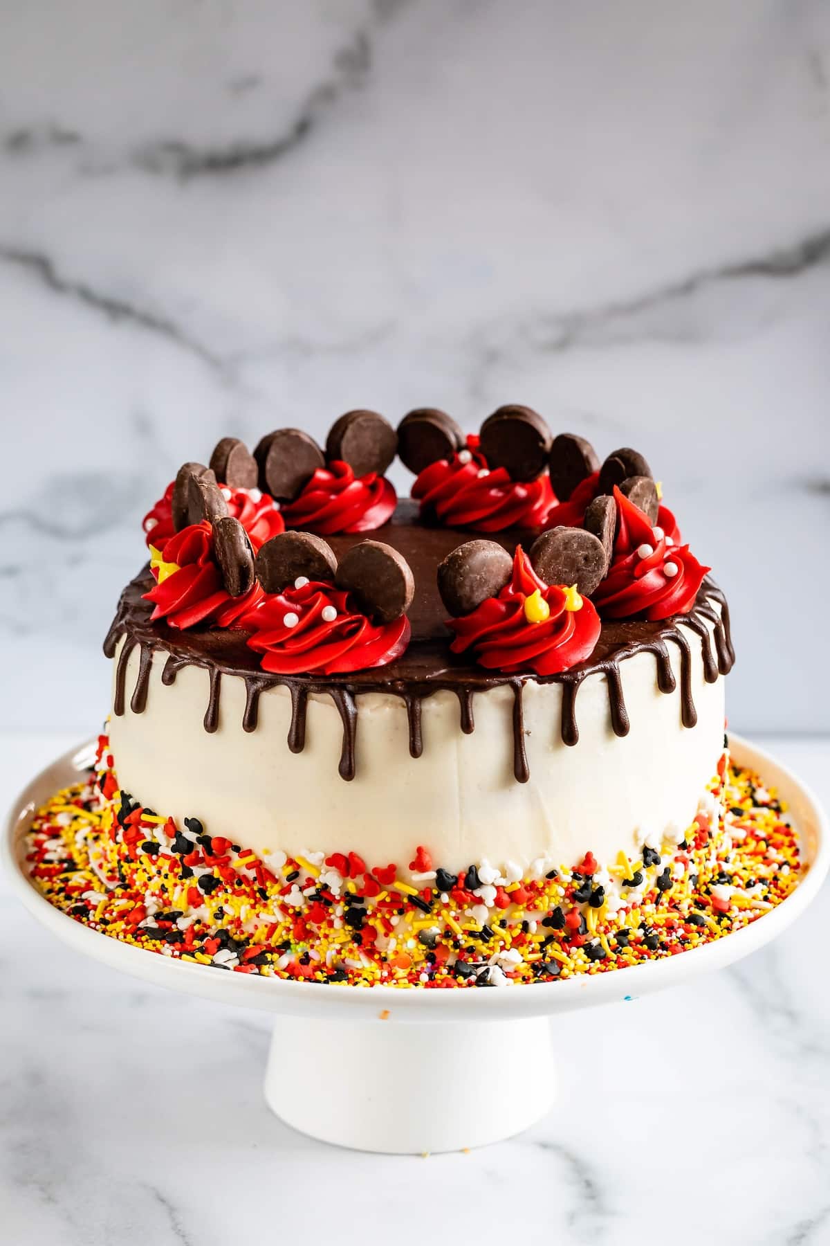 A white frosted cake with a chocolate drip, decorated with red and yellow icing, chocolate pieces, and colorful sprinkles on the base, displayed on a white cake stand against a marble background.