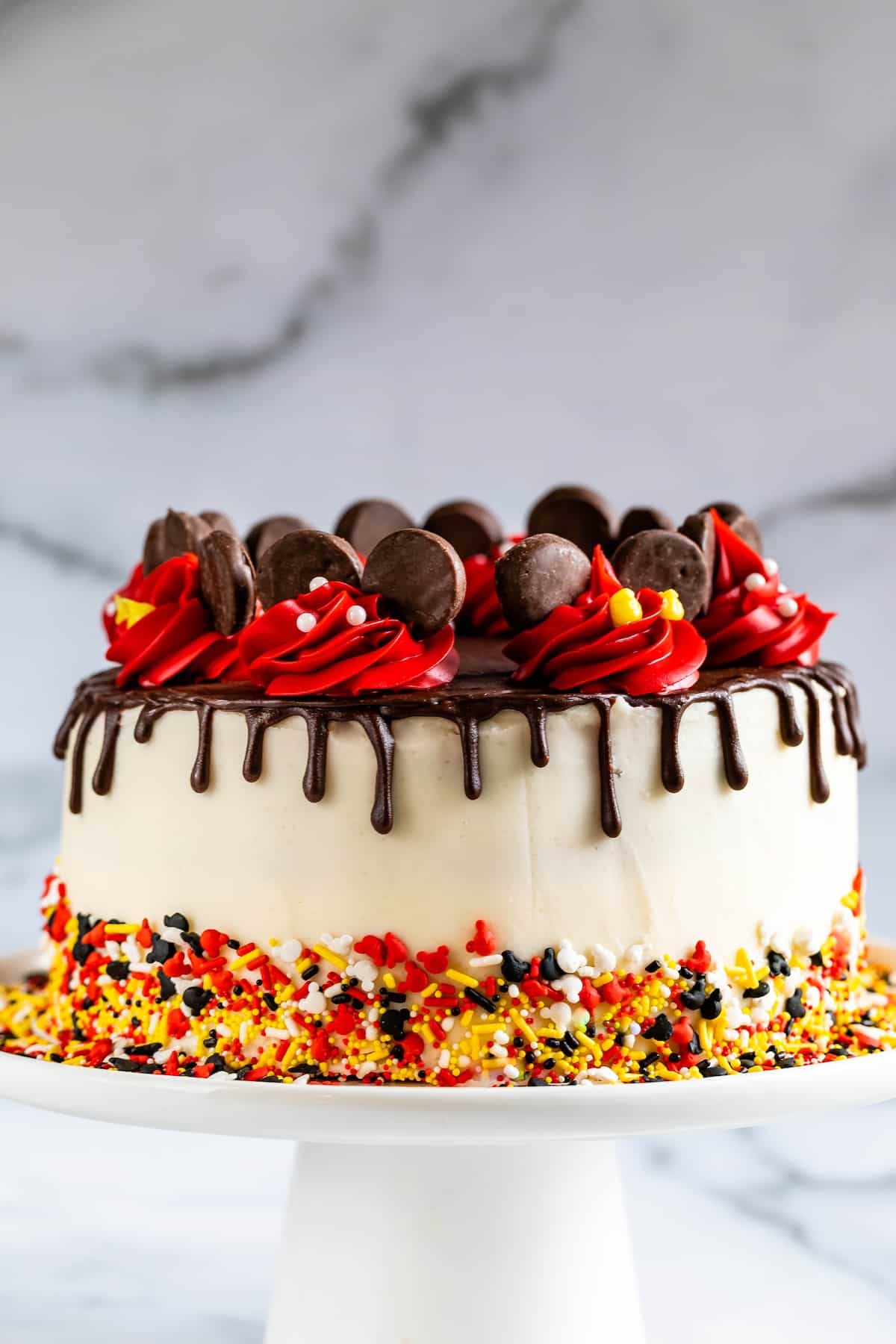 A round cake with white frosting, chocolate ganache drips, red swirled icing, chocolate cookies on top, and colorful sprinkles around the base, displayed on a white cake stand.