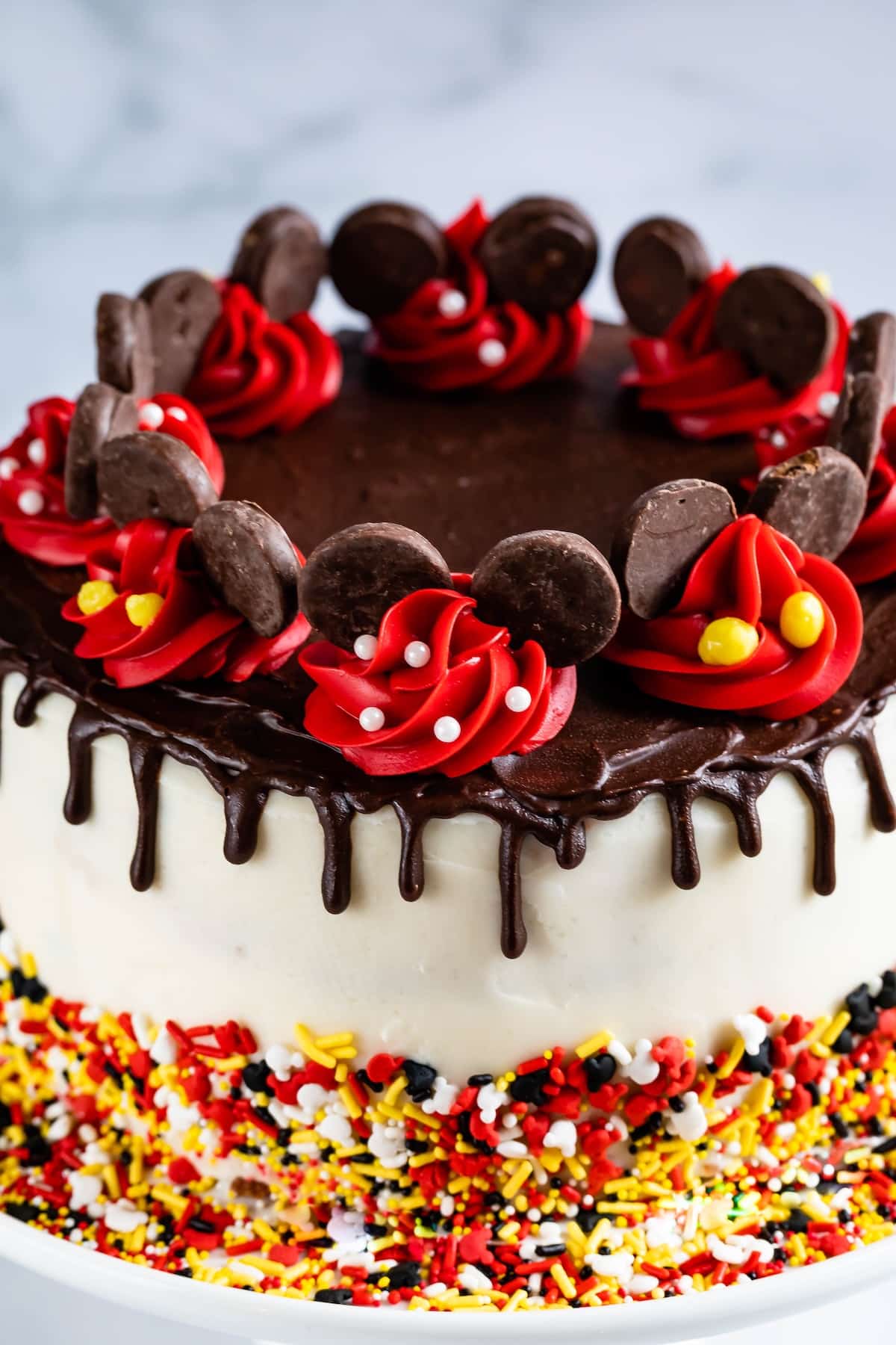 A round cake with white frosting, a chocolate drip topping, and colorful sprinkles at the base. The top is decorated with red icing swirls, chocolate cookies, and edible pearls, resembling a Mickey Mouse theme.