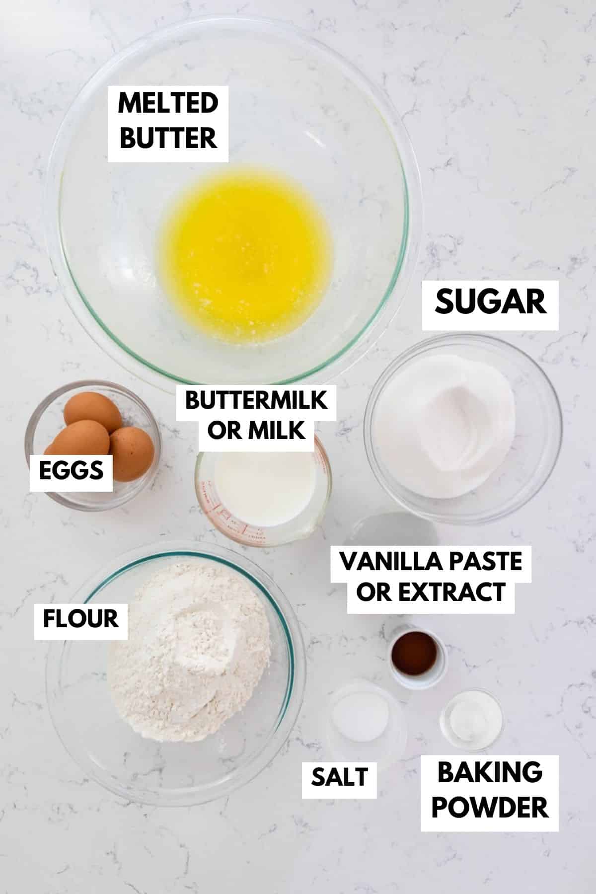 Ingredients for a recipe arranged on a marble surface, including bowls of melted butter, sugar, and flour, plus eggs, buttermilk or milk, vanilla paste or extract, salt, and baking powder. Each item is labeled in bold text.