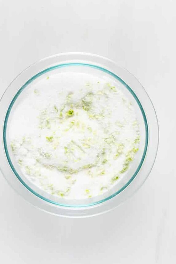 A bowl filled with white sugar mixed with small green flecks of lime zest, sitting on a light gray surface.