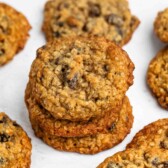 A stack of oatmeal cookies with chocolate chips is surrounded by more cookies on a white surface. The cookies have a golden-brown color and a textured surface.