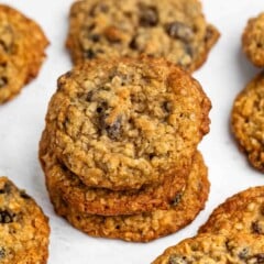 A stack of oatmeal cookies with chocolate chips is surrounded by more cookies on a white surface. The cookies have a golden-brown color and a textured surface.