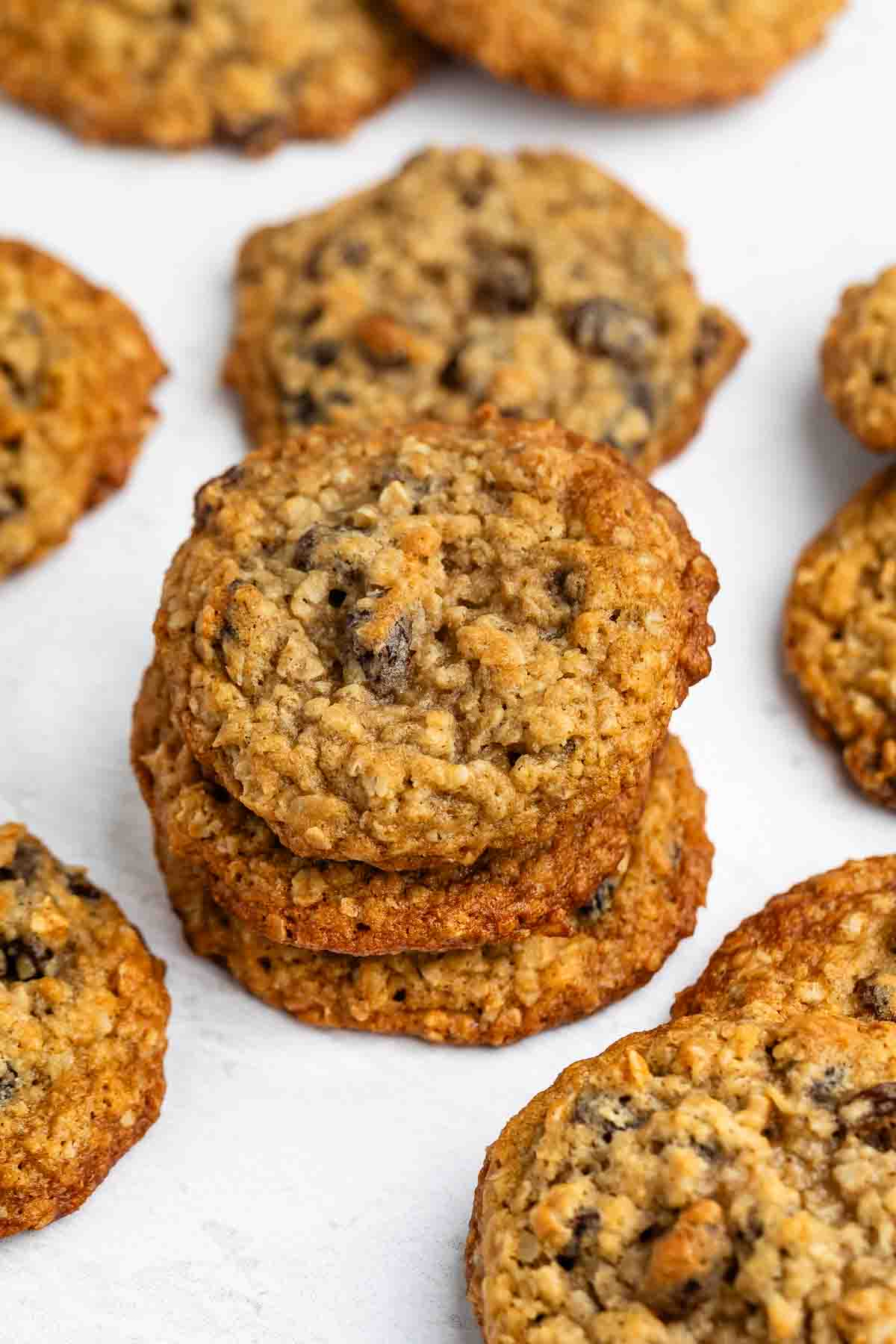 A stack of oatmeal cookies with chocolate chips is surrounded by more cookies on a white surface. The cookies have a golden-brown color and a textured surface.