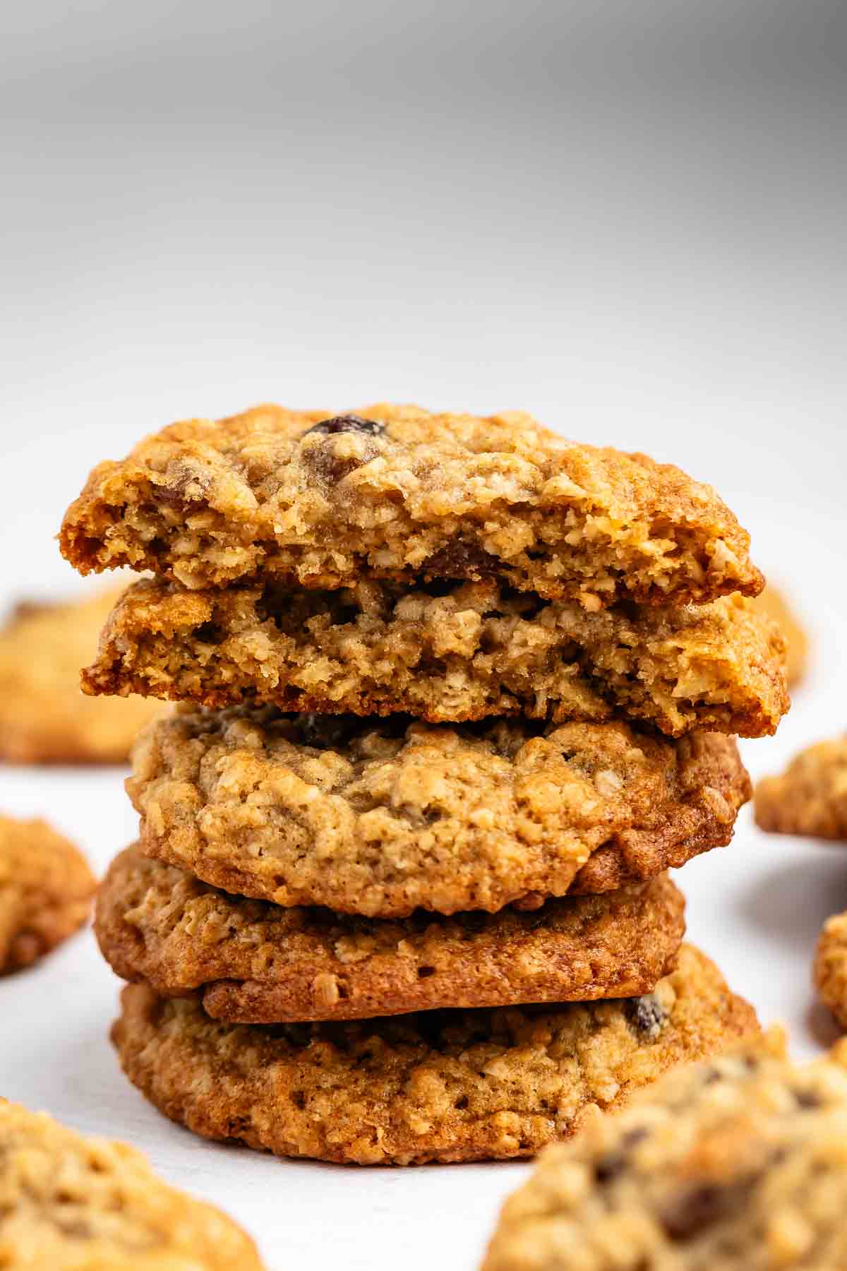 A stack of oatmeal cookies, with the top one broken in half to reveal a chewy interior. Other cookies are scattered in the background on a light surface.