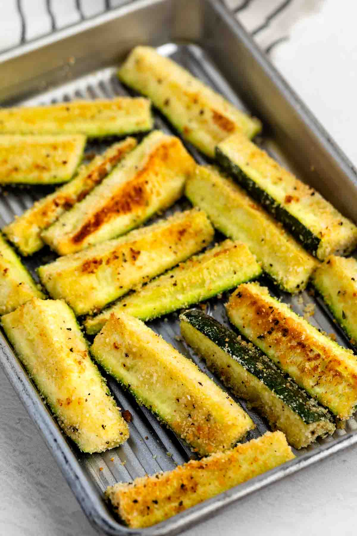 A baking tray filled with golden-brown, breaded zucchini sticks arranged in rows, showing a crispy texture and seasoning on the surface.