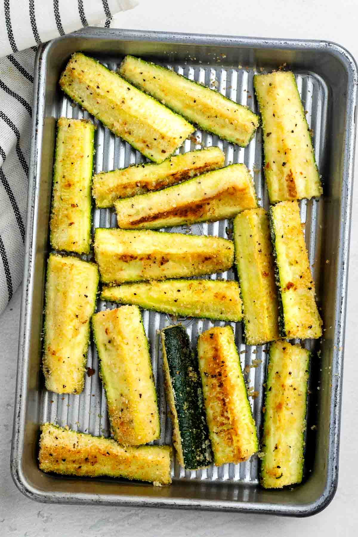 A baking tray filled with golden-brown roasted zucchini sticks, seasoned with herbs and spices. A striped kitchen towel is partially visible on the left side.