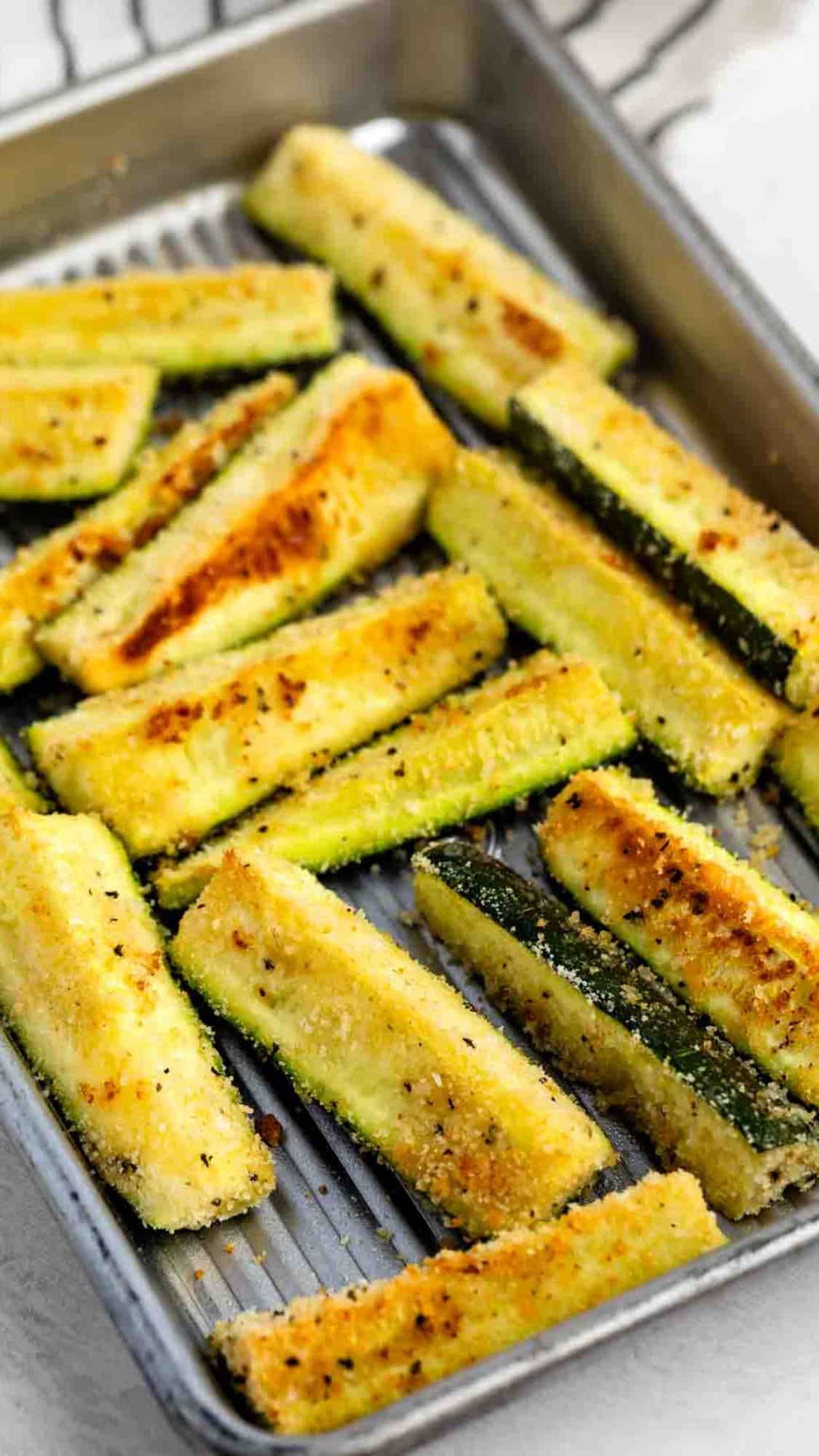 Golden, crispy zucchini sticks coated with breadcrumbs and seasoning are arranged in a single layer on a metal baking tray. The sticks are baked and slightly browned on top.