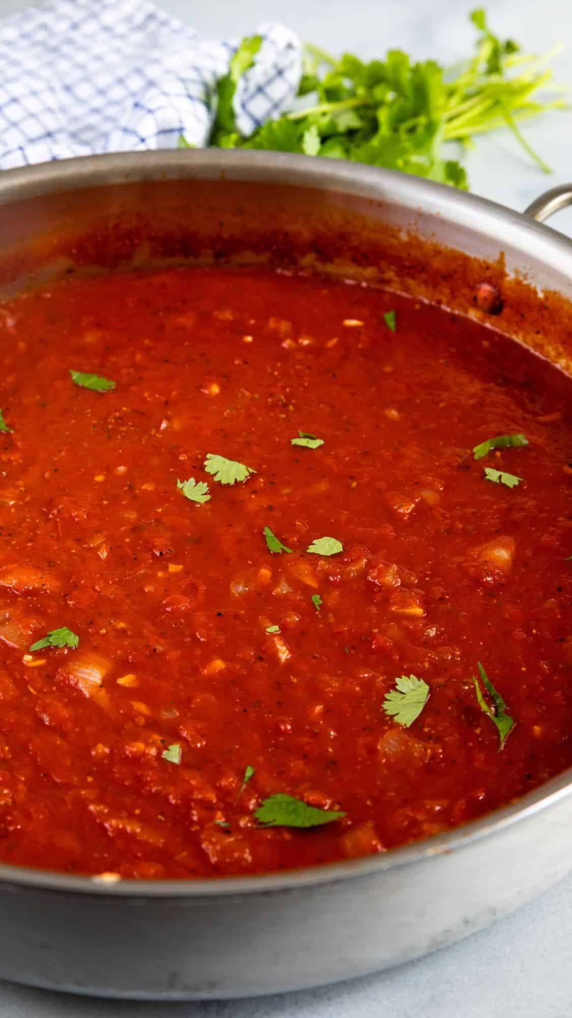 A stainless steel pan filled with chunky red tomato sauce, garnished with fresh cilantro leaves. In the background, a checkered cloth and a small bunch of herbs are visible on a light surface.