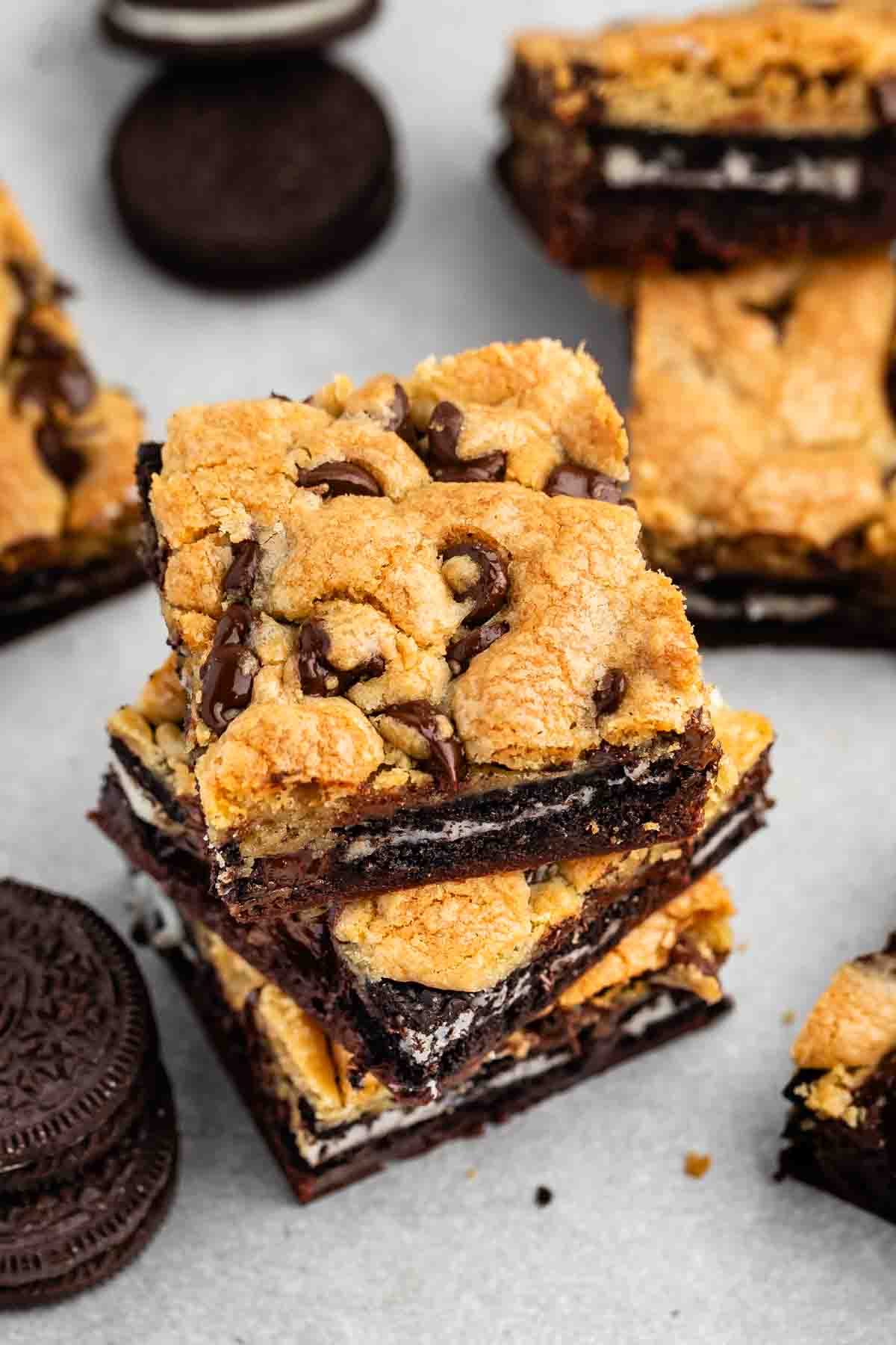 A close-up of dessert bars stacked on a surface, featuring layers of chocolate chip cookie, Oreo cookies, and brownies. Other Oreo cookies and dessert bars are visible in the background.