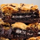 A close-up of stacked brookies—also known as slutty brownies—showing layers of chocolate chip cookie, Oreo cookies, and fudgy brownie. The treats look gooey and rich, with melted chocolate chips on top.