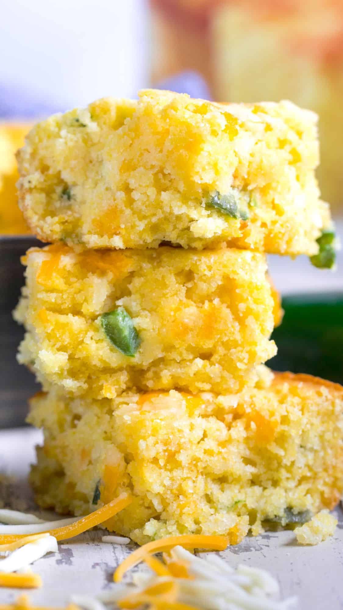 Two stacked pieces of cornbread with visible cheese and chopped green peppers. The texture looks soft and crumbly, with some shredded cheese scattered around. The background is blurred, focusing on the cornbread.