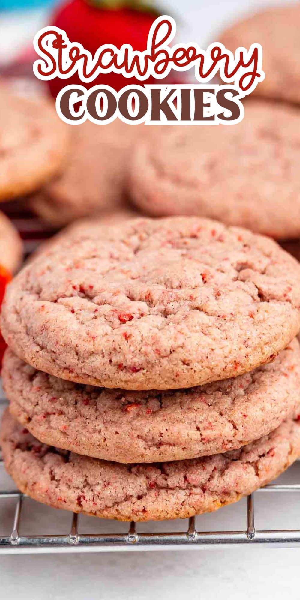 A close-up of three stacked strawberry cookies with visible red strawberry bits, resting on a wire cooling rack, with more cookies and part of a strawberry in the background. Text at the top reads Strawberry Cookies.