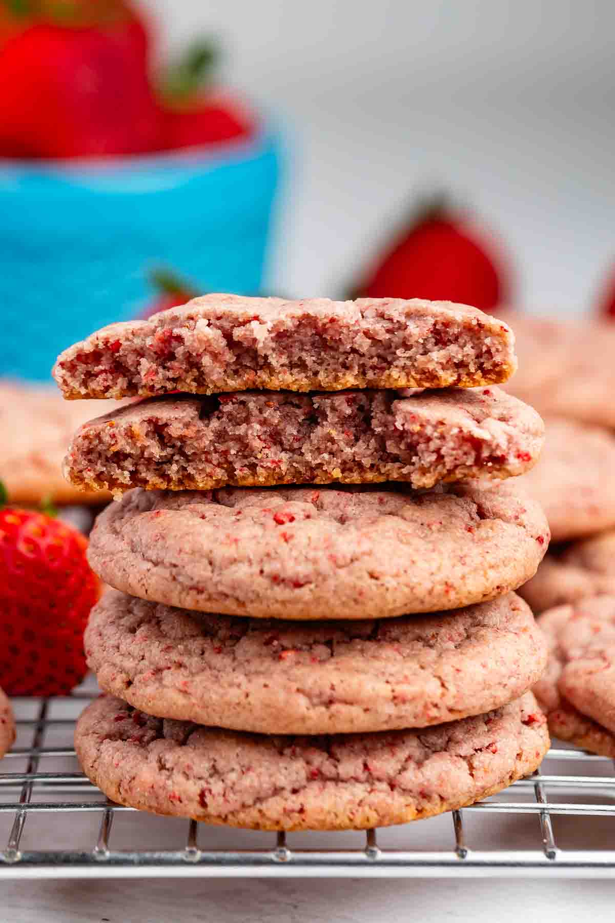 A close-up of three stacked strawberry cookies with visible red strawberry bits, resting on a wire cooling rack, with more cookies and part of a strawberry in the background.