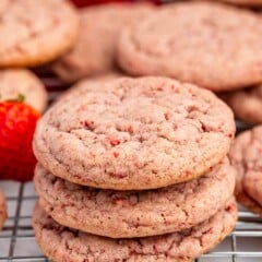 A close-up of three stacked strawberry cookies with visible red strawberry bits, resting on a wire cooling rack, with more cookies and part of a strawberry in the background.