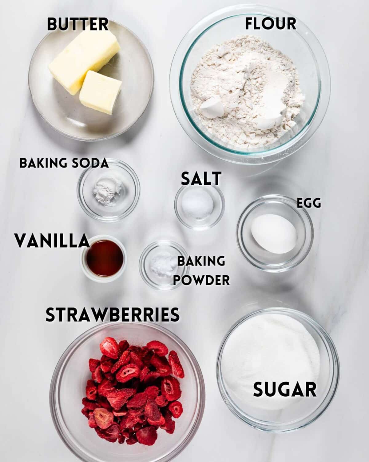Overhead view of baking ingredients in bowls, labeled: butter, flour, baking soda, salt, egg, vanilla, baking powder, strawberries, and sugar, arranged on a white surface.