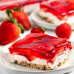A dessert on a white plate features a layered strawberry pretzel salad. It includes a pretzel crust, cream cheese filling, and strawberry gelatin with fresh strawberry slices on top. Fresh strawberries decorate the surrounding area.