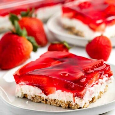 A dessert on a white plate features a layered strawberry pretzel salad. It includes a pretzel crust, cream cheese filling, and strawberry gelatin with fresh strawberry slices on top. Fresh strawberries decorate the surrounding area.