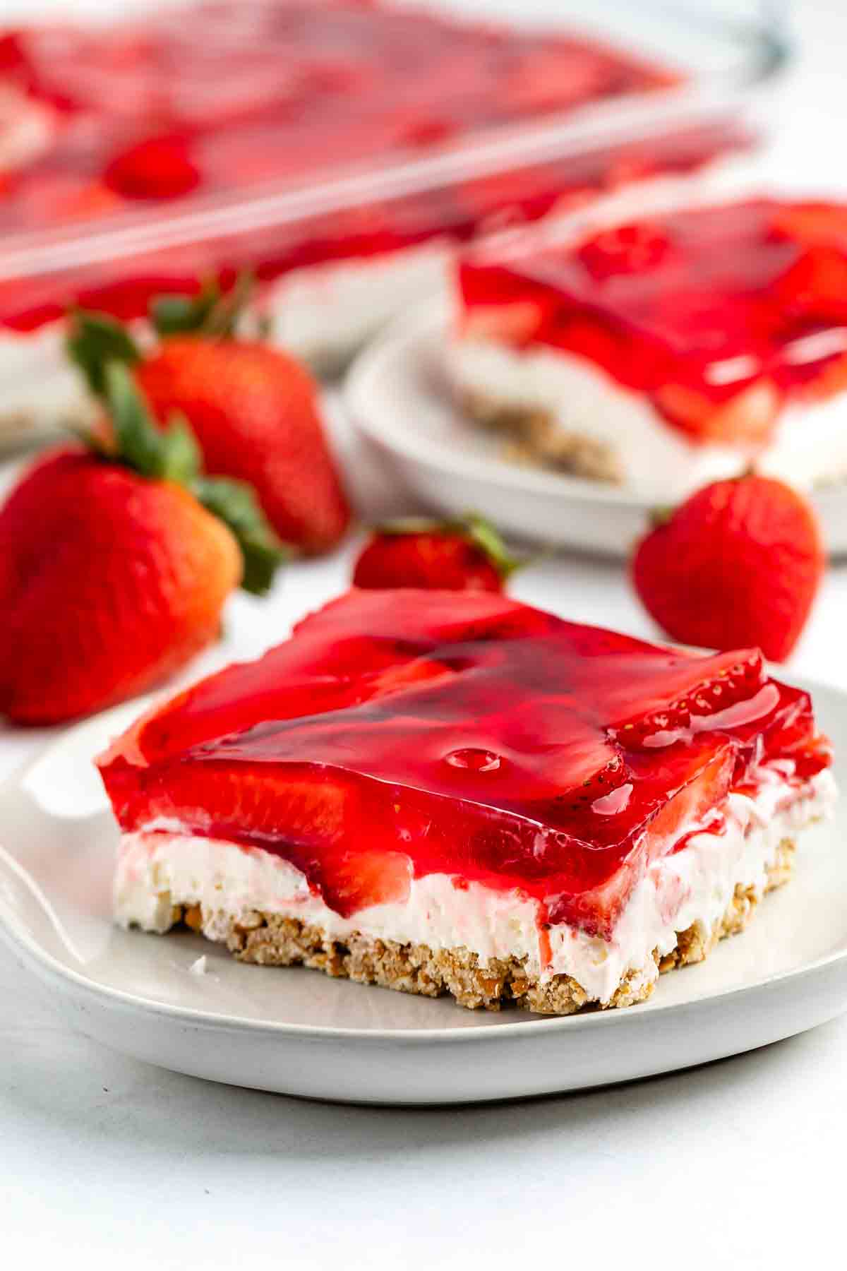 A dessert on a white plate features a layered strawberry pretzel salad. It includes a pretzel crust, cream cheese filling, and strawberry gelatin with fresh strawberry slices on top. Fresh strawberries decorate the surrounding area.