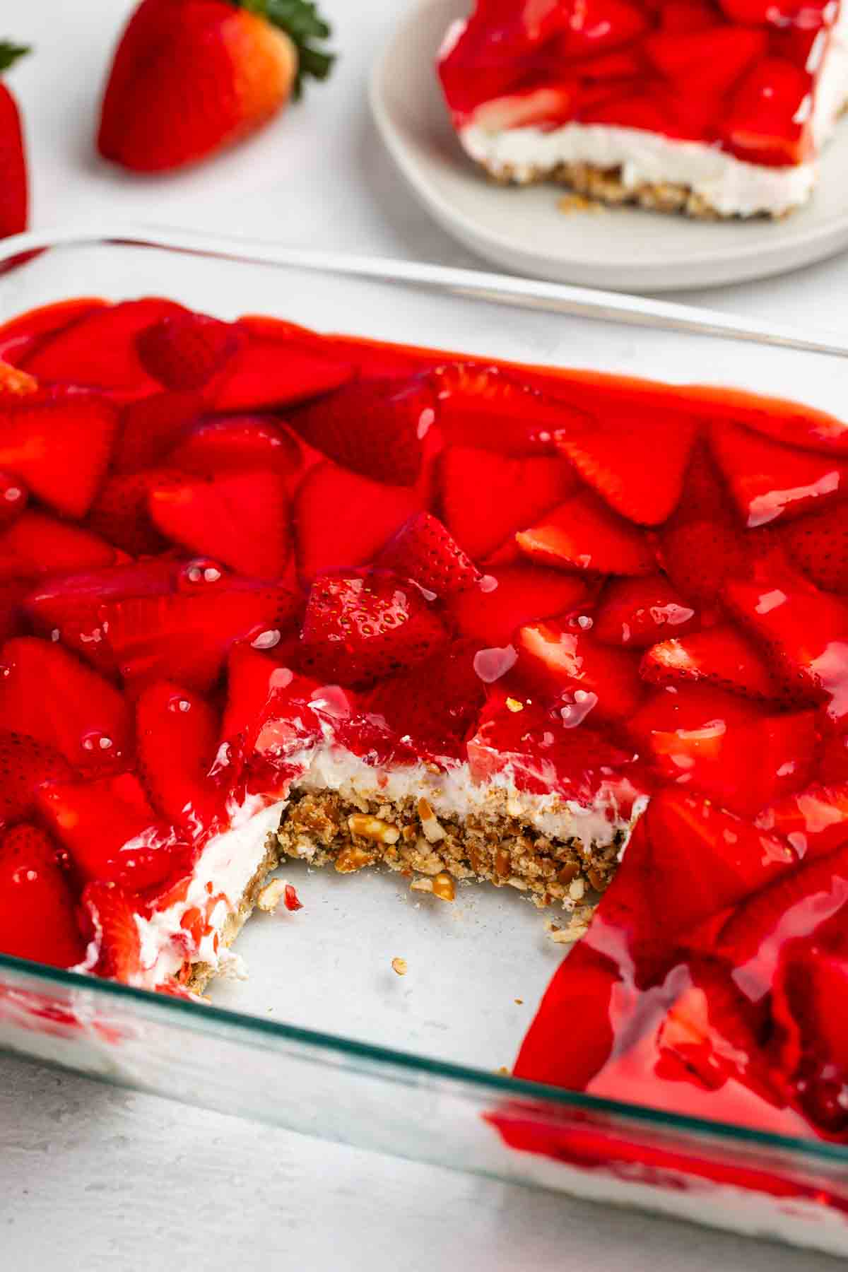 A glass dish with a dessert featuring layers of pretzel crust, creamy filling, and strawberry gelatin topping. A slice is missing, revealing the layers. Fresh strawberries are scattered on top and nearby.