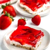 A slice of strawberry pretzel dessert on a white plate. It has a layered structure with a pretzel crust, creamy filling, and strawberry gelatin topping. Fresh strawberries surround the plate.
