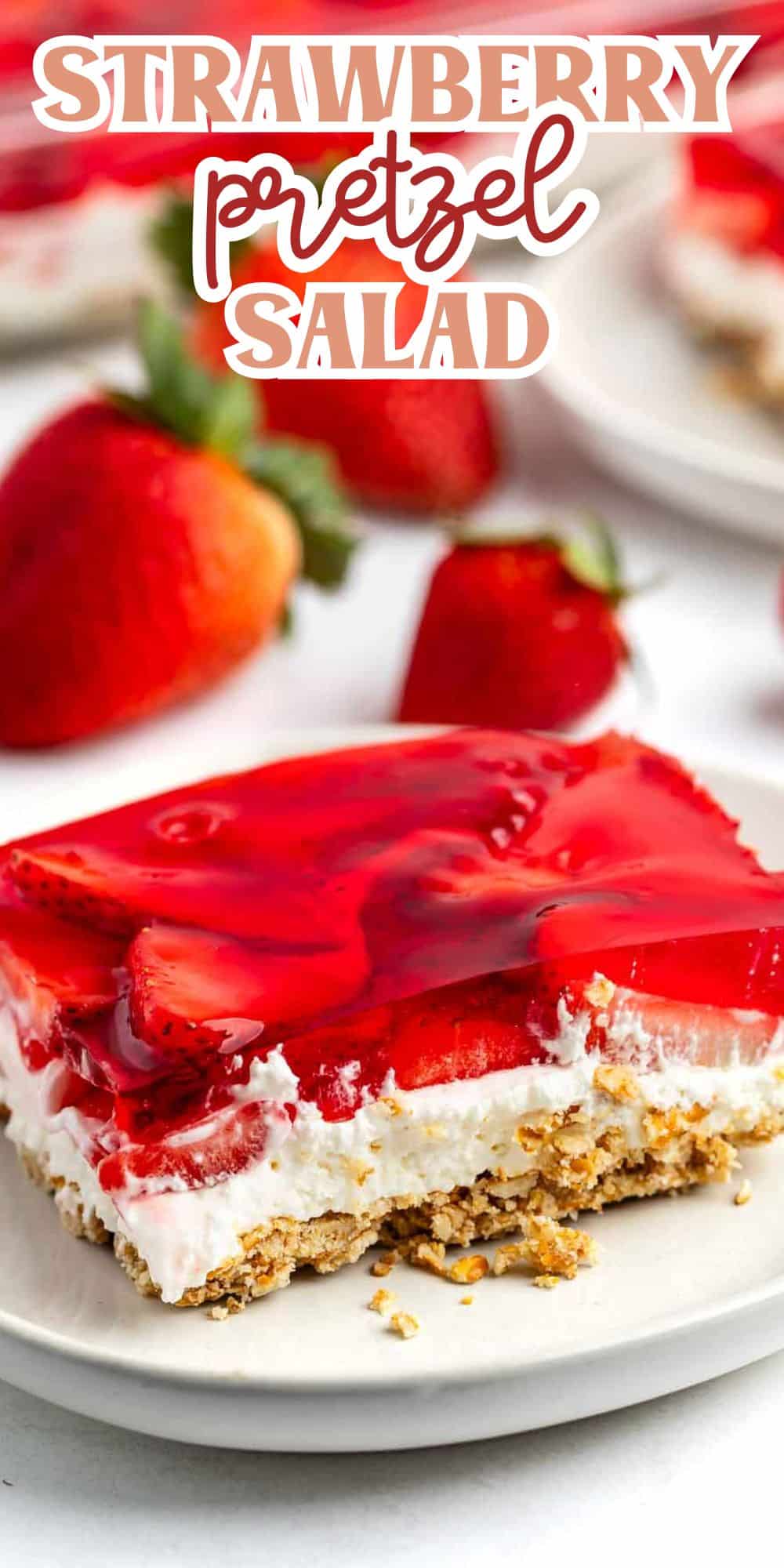 A slice of strawberry pretzel salad on a white plate, topped with strawberry pieces and red gelatin, with a crushed pretzel crust and creamy filling. Fresh strawberries in the background. Text at the top reads Strawberry Pretzel Salad.