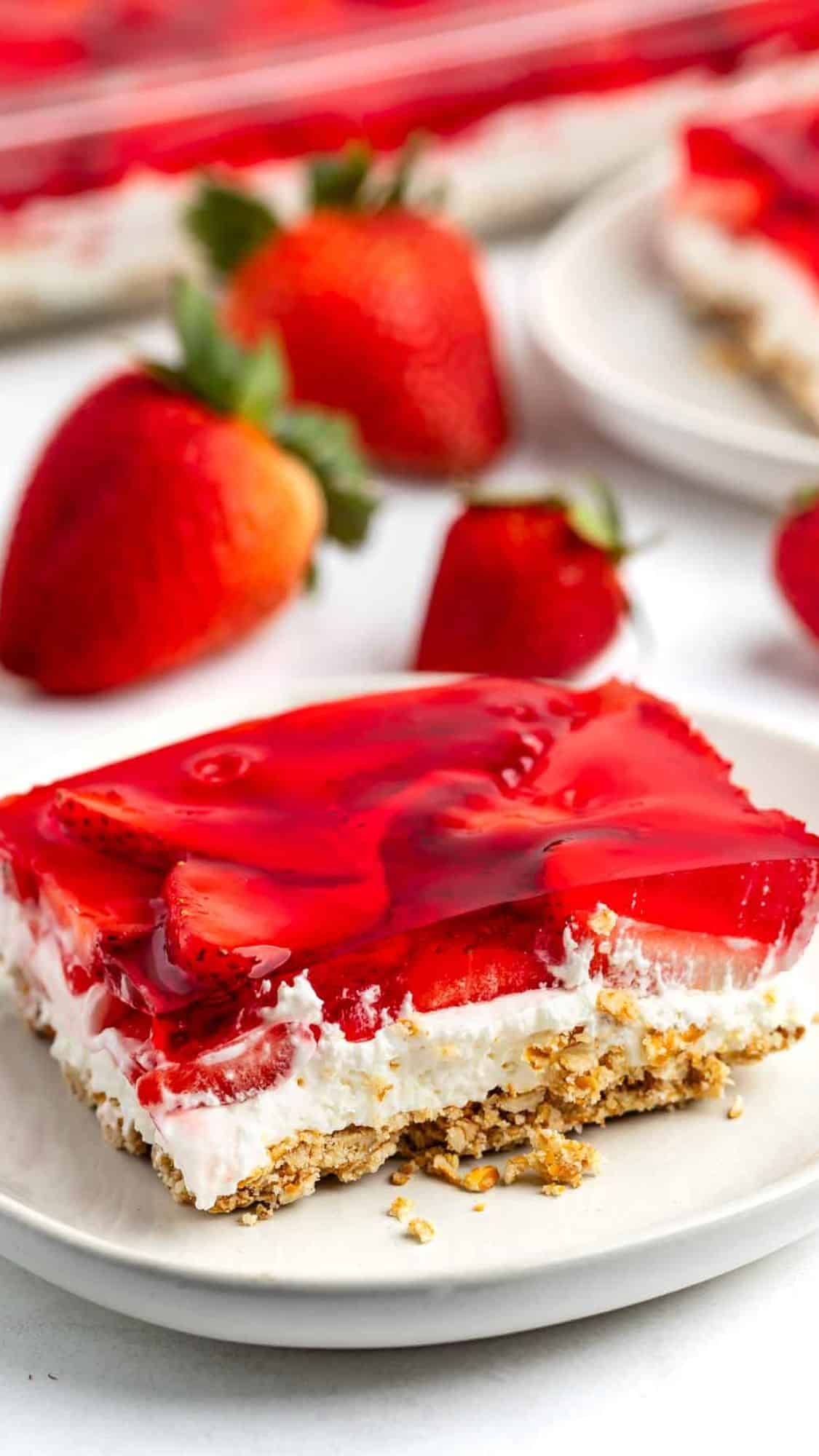A dessert bar on a plate with a crumbly base, layered with white cream, topped with sliced strawberries and a red gelatin layer. Whole strawberries are in the background, and another plate is partially visible.