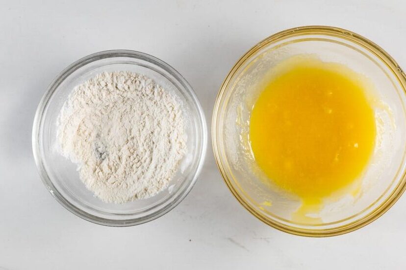 Two glass bowls are on a white surface. The left bowl contains a beaten yellow egg mixture, and the right bowl is filled with white flour.