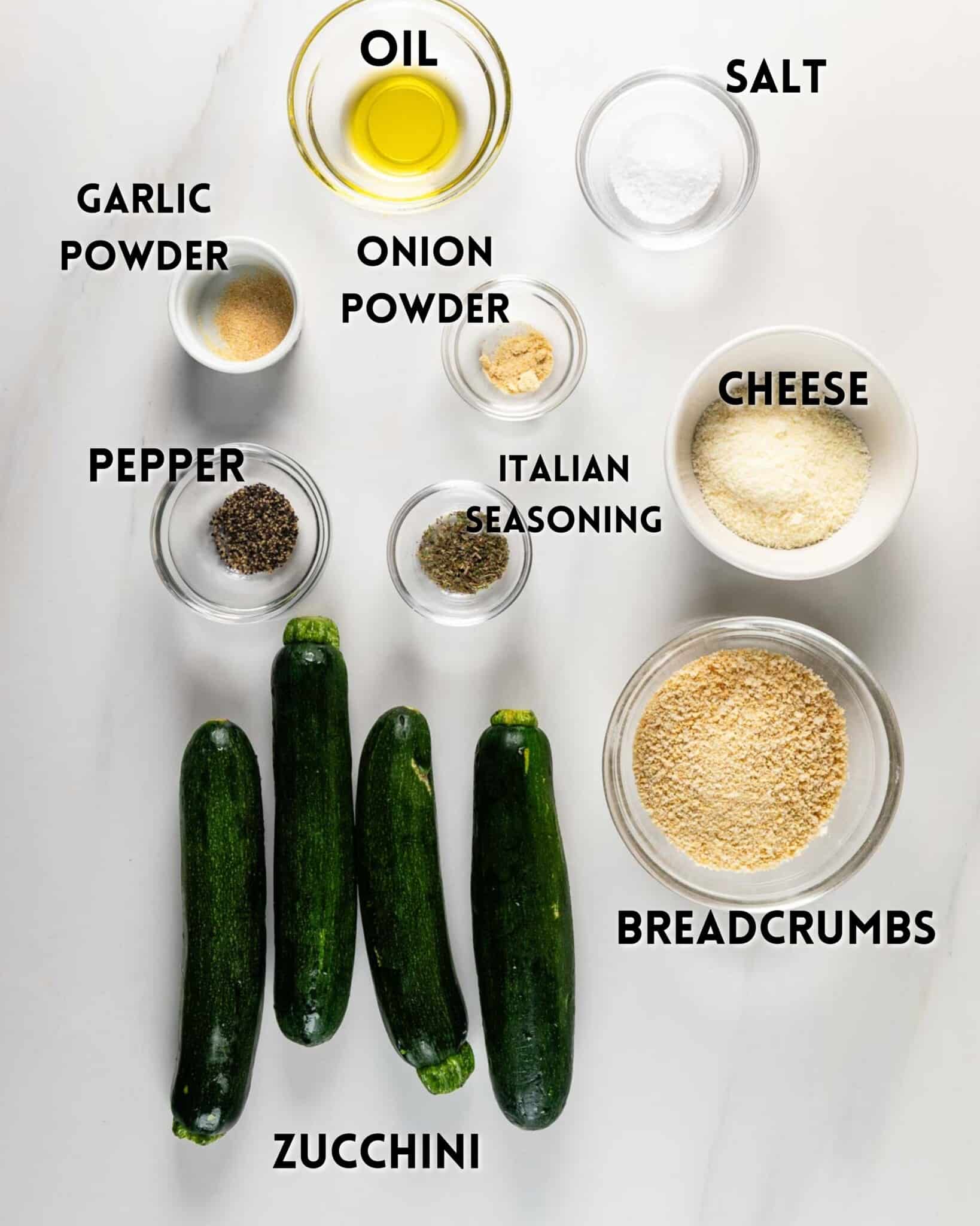 Four zucchinis, small bowls of oil, salt, garlic powder, onion powder, pepper, Italian seasoning, cheese, and breadcrumbs arranged on a white surface, each ingredient labeled with text.