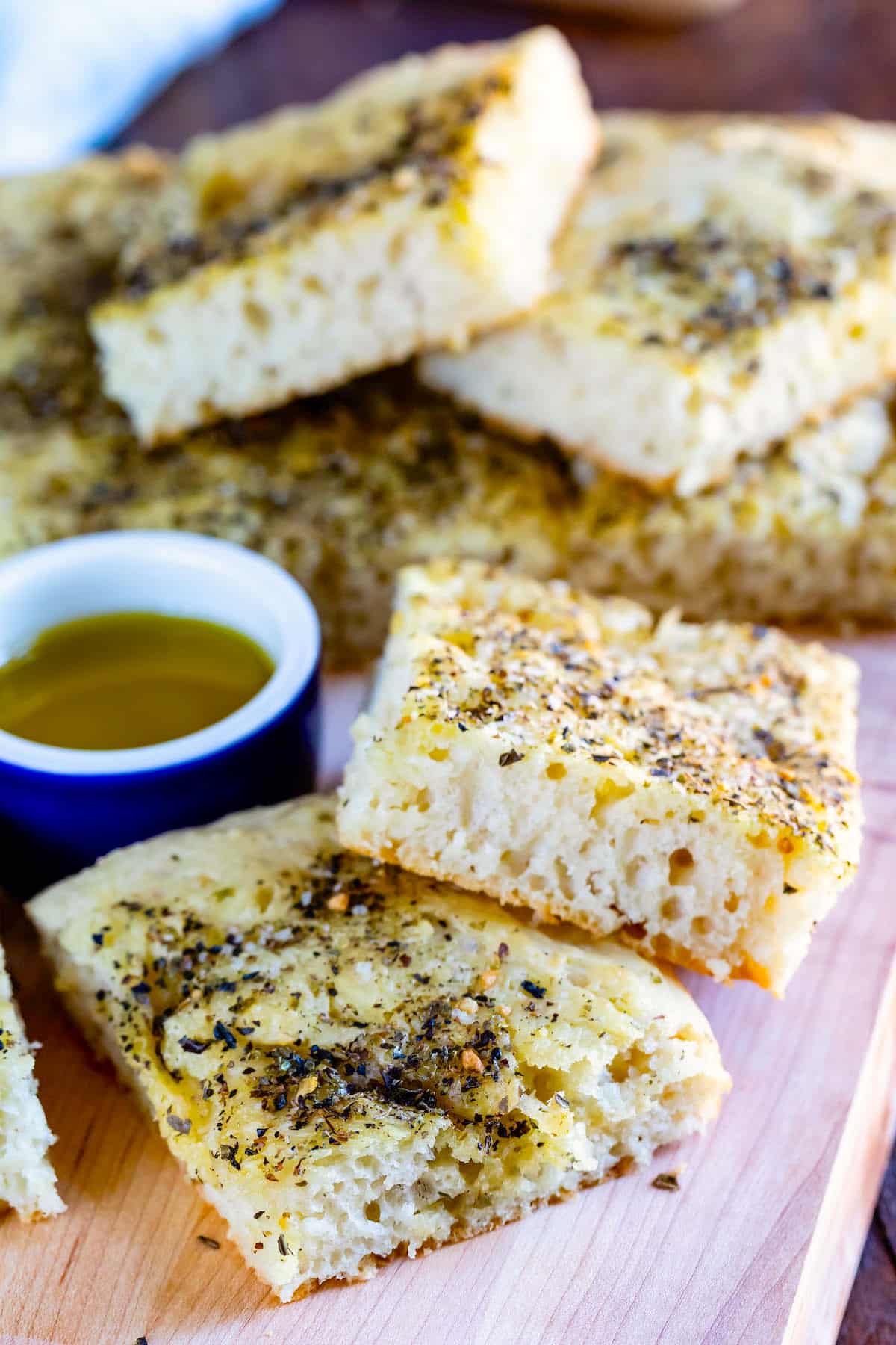 Slices of fluffy focaccia bread topped with herbs are arranged on a wooden board, with a small bowl of olive oil next to them for dipping.