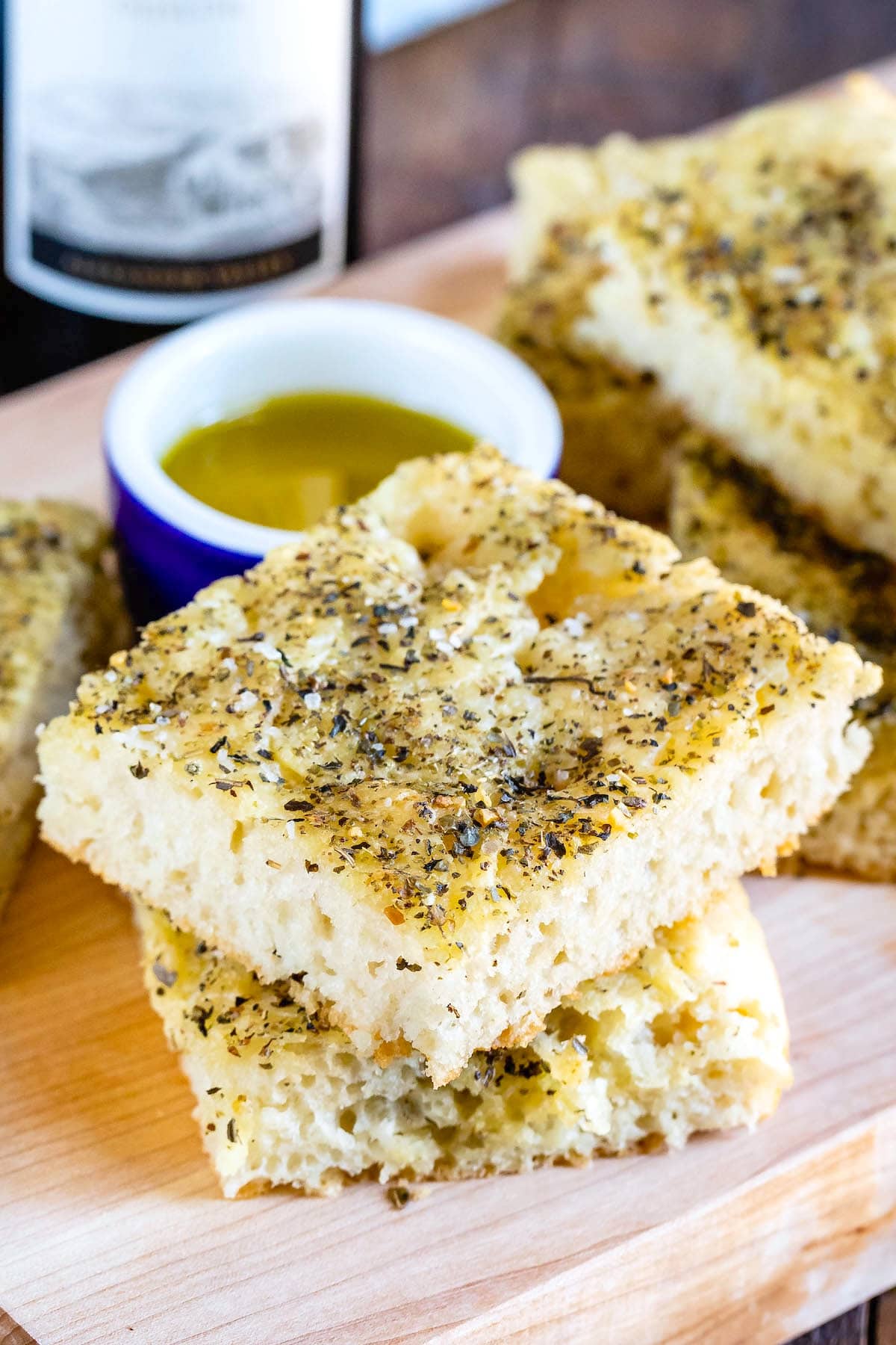Two thick slices of focaccia bread stacked on a wooden board, topped with herbs and served with a small dish of olive oil for dipping. A bottle of wine is partially visible in the background.