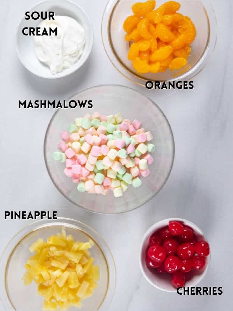 Five bowls on a white surface hold ingredients for an ambrosia salad recipe: sour cream, mandarin orange segments, colorful mini marshmallows, pineapple chunks, and red cherries. Each bowl is neatly labeled with its contents.
