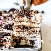 A hand lifts a slice of layered ice cream cake topped with Oreo cookies and colorful sprinkles from a baking pan, showing visible layers of cookies and cream inside.
