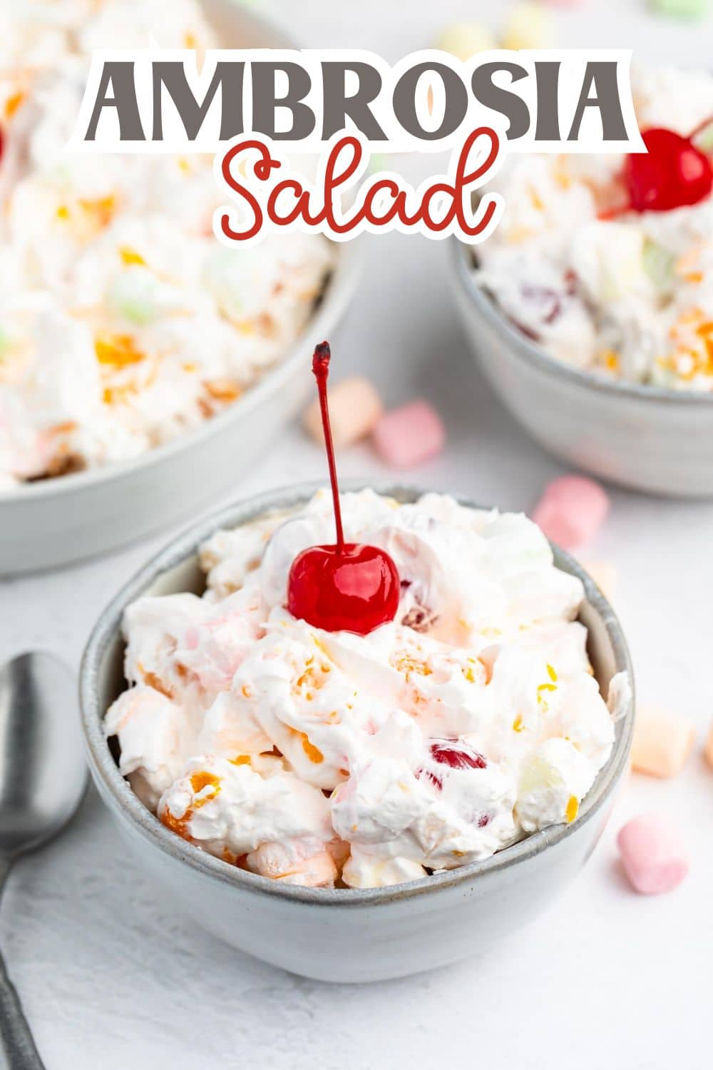 A bowl of ambrosia salad topped with a cherry, surrounded by pastel mini marshmallows, with the words Ambrosia Salad written above in bold letters. Two more bowls of salad are blurred in the background.