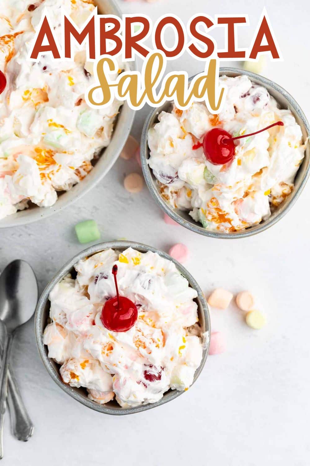 Two bowls and one large dish filled with Ambrosia salad topped with cherries. Colorful mini marshmallows are scattered nearby, and two spoons are set to the side. The text AMBROSIA Salad is at the top.