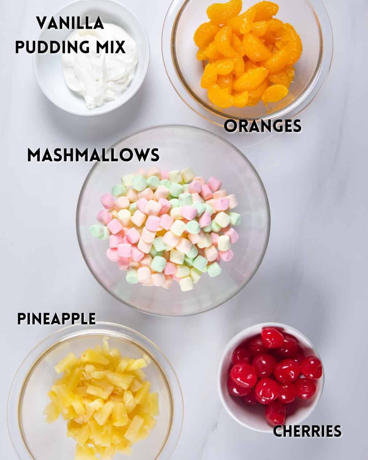 Five bowls with labeled ingredients: vanilla pudding mix, mandarin orange segments, colorful mini marshmallows, pineapple chunks, and maraschino cherries, arranged on a white surface.