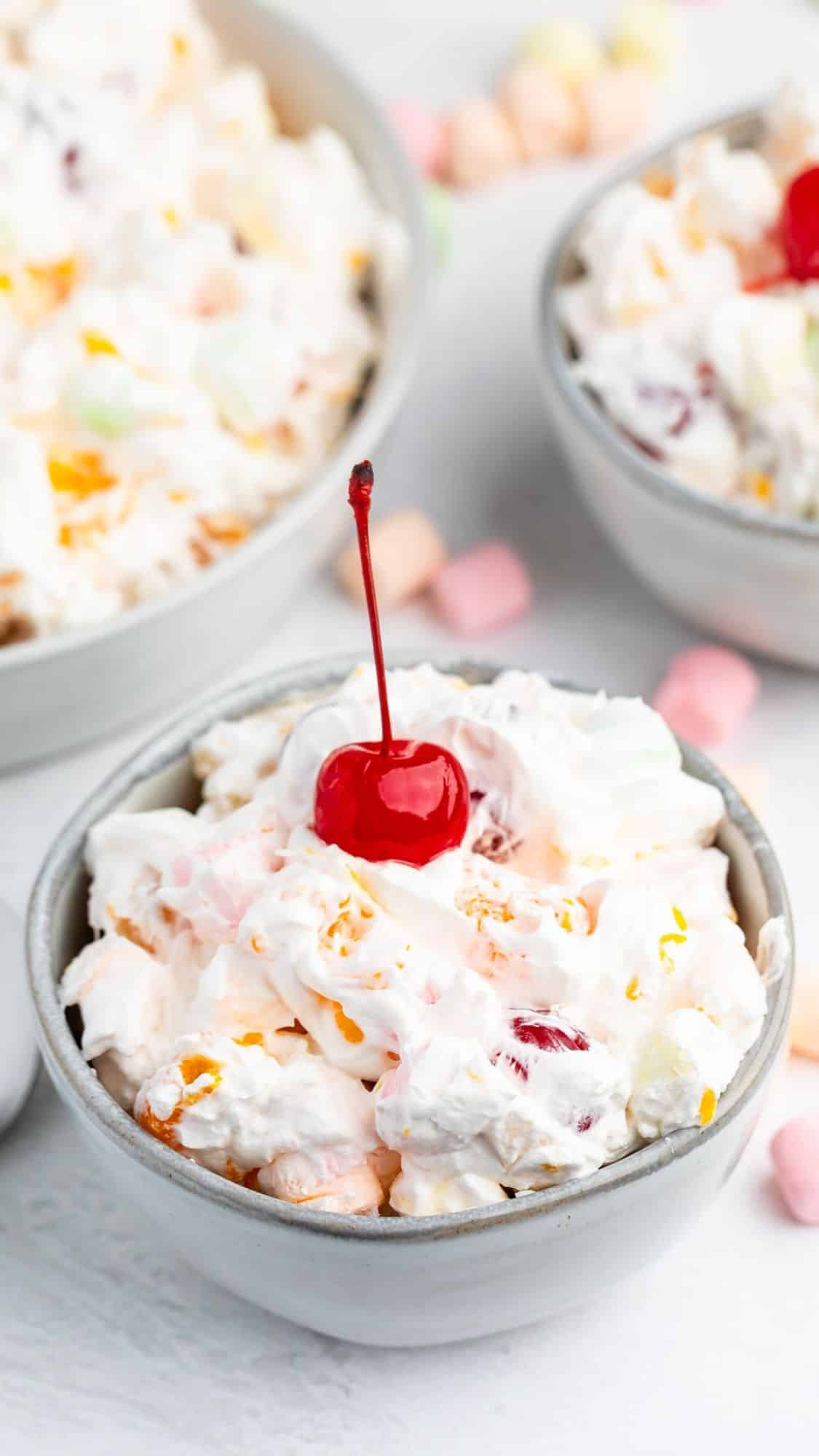 A bowl of creamy fruit salad topped with a bright red maraschino cherry, surrounded by other bowls of fruit salad and pastel mini marshmallows in the background.