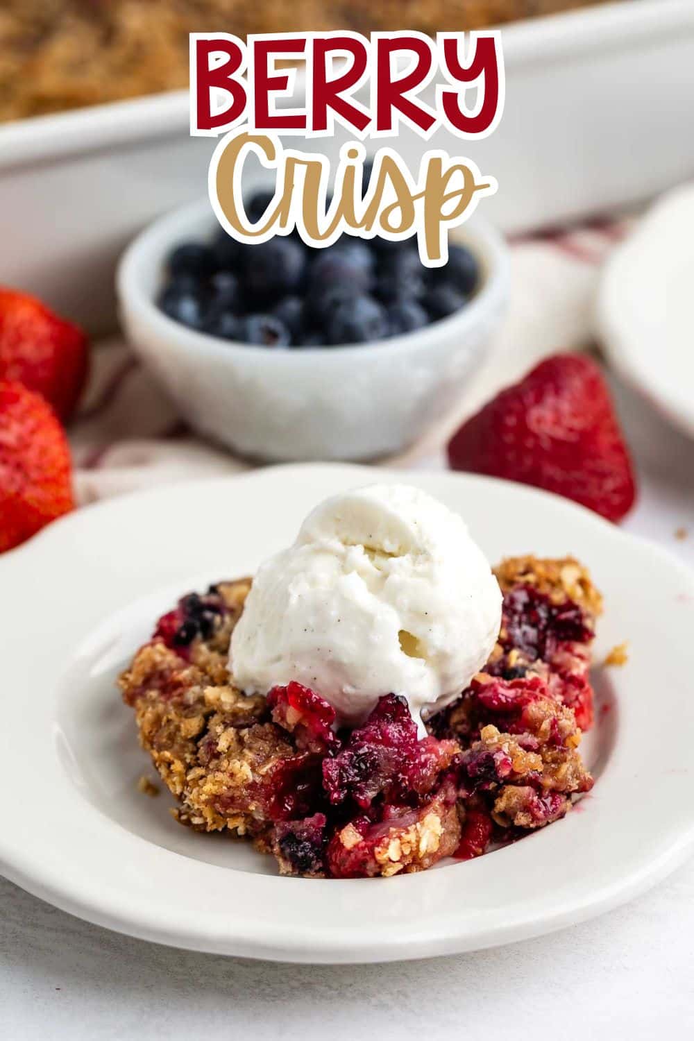 A serving of berry crisp topped with a scoop of vanilla ice cream sits on a white plate, with fresh strawberries and a bowl of blueberries in the background. Text at the top reads Berry Crisp.