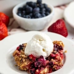 A serving of berry crumble topped with a scoop of vanilla ice cream on a white plate, with fresh strawberries and a bowl of blueberries in the background.