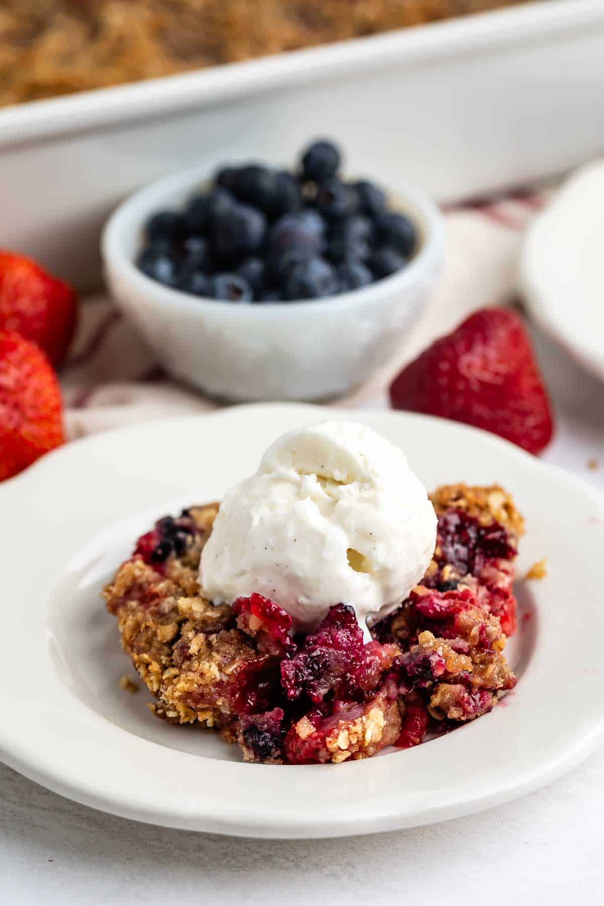 A serving of berry crumble topped with a scoop of vanilla ice cream on a white plate, with fresh strawberries and a bowl of blueberries in the background.