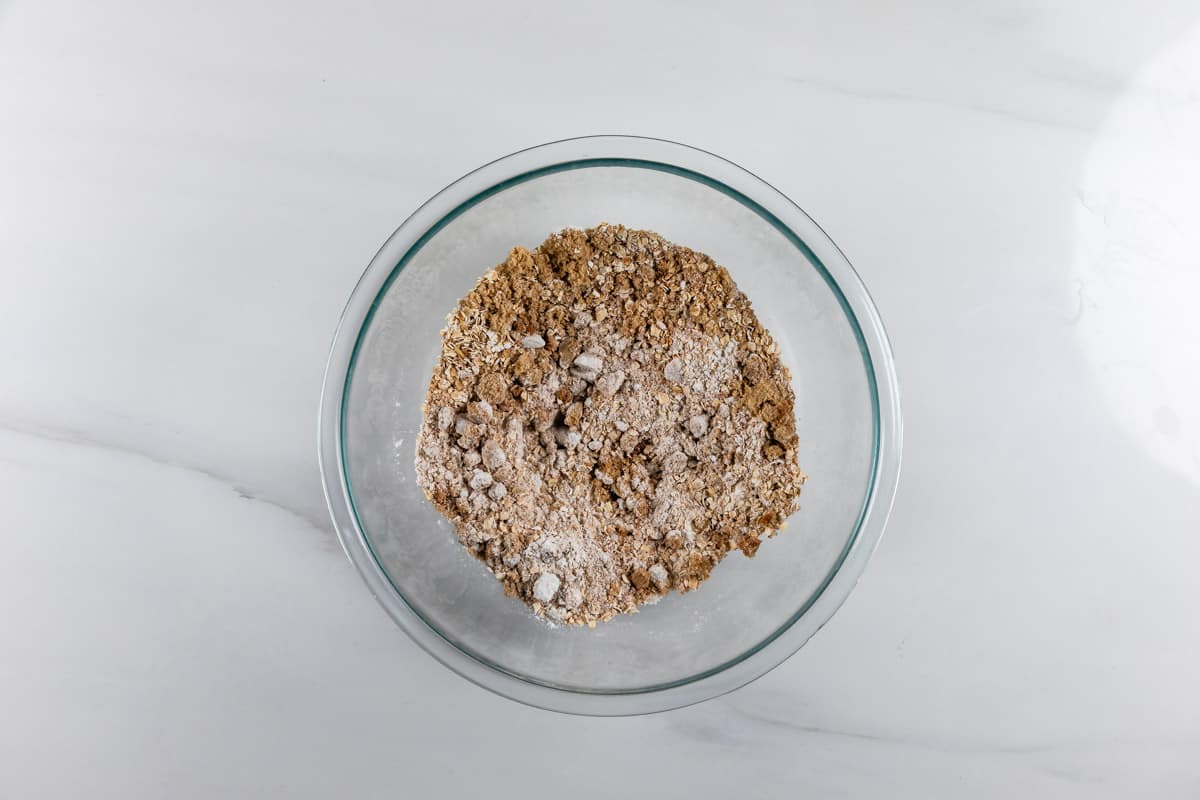 A glass bowl filled with a mixture of dry baking ingredients, including flour, oats, and brown sugar, sitting on a white marble surface.