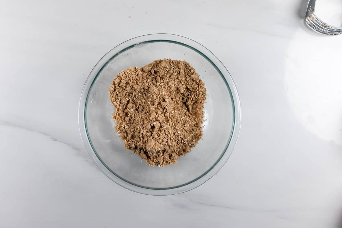 A glass bowl filled with a crumbly brown mixture sits on a white marble surface, with part of a metal pastry cutter visible in the top right corner.