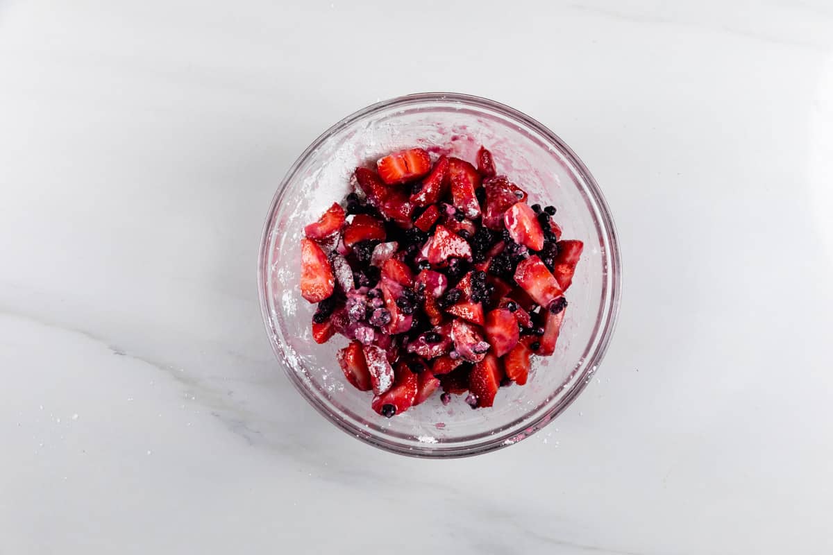 A glass bowl filled with chopped strawberries and blueberries coated in powdered sugar, placed on a white marble surface.