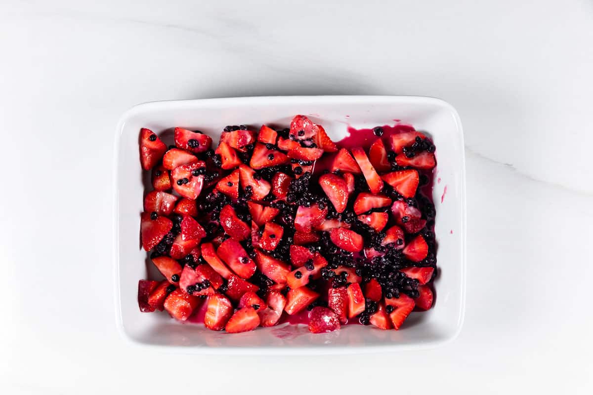 A white rectangular dish filled with chopped strawberries and blueberries mixed together, with some berry juice visible on the fruits and dish, set on a white surface.