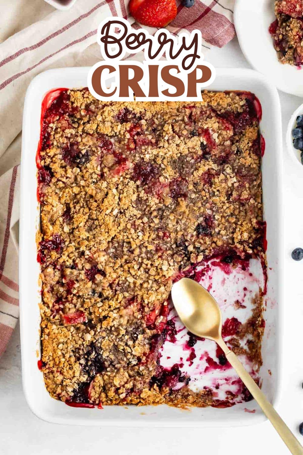A rectangular baking dish filled with berry crisp, topped with a golden crumb layer. A serving has been scooped out, revealing juicy berries underneath. A gold spoon rests in the dish, and Berry Crisp is written at the top.